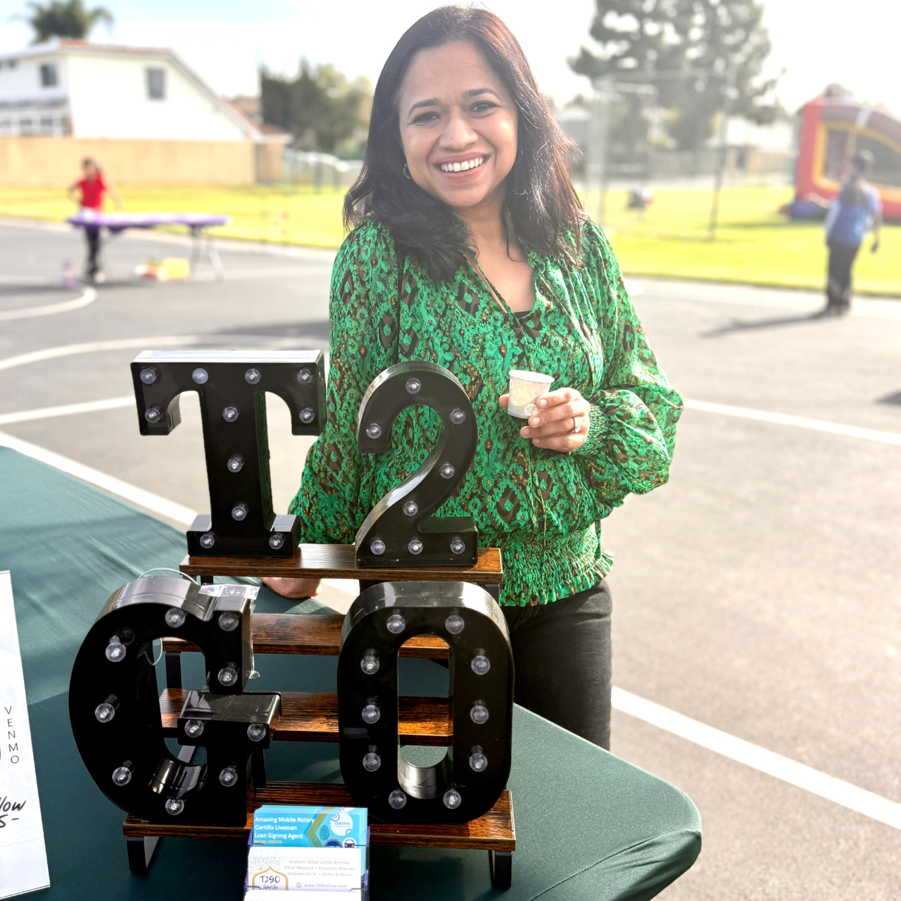 Woman in green patterned shirt holding a cup, standing next to decorative letters on a table outdoors.